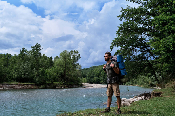 Νέος όμορφος Καυκάσιος άνδρας με γενειάδα και dreadlocks περπατά κατά μήκος όχθης του ποταμού βουνού με μεγάλο σακίδιο πεζοπορίας και απολαμβάνοντας θέα και ομορφιές. Guy είναι ταξιδιώτης και τουρίστας. - Φωτογραφία, εικόνα