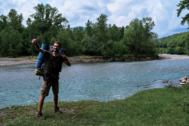 Handsome Caucasian man with beard and dreadlocks stands on bank of mountain river with large hiking backpack and takes selfie on his phone. Guy runs travel blog. - Foto, Imagem