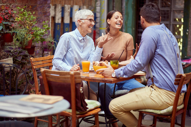 elderly woman  having conversation with young adult female and her boyfriend, smiling, having breakfast together in outdoor cafe. family communication  concept. - Photo, image