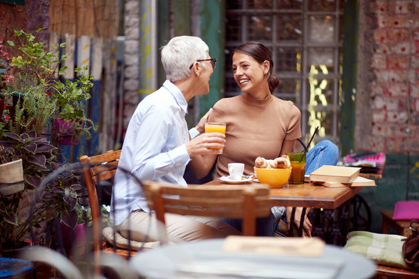 elderly woman and young adult female having breakfast together, looking each other, smiling. mother and daughter concept. - Photo, image