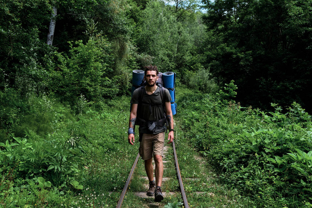 Νεαρός όμορφος Καυκάσιος άνδρας με γένια και ράστα περπατά κατά μήκος του σιδηροδρόμου ανάμεσα στο πράσινο δάσος. Ταξιδιώτης με μεγάλο σακίδιο περπατά κατά μήκος των σιδηροτροχιών και κοιμάται μπροστά. - Φωτογραφία, εικόνα