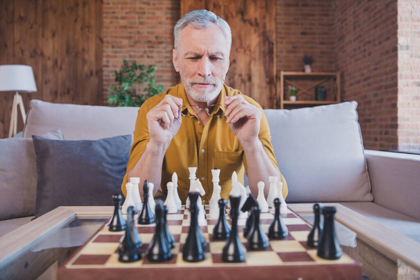 Photo of smart thinking mature man hold spectacles play chess wear yellow shirt at home alone - Foto, imagen