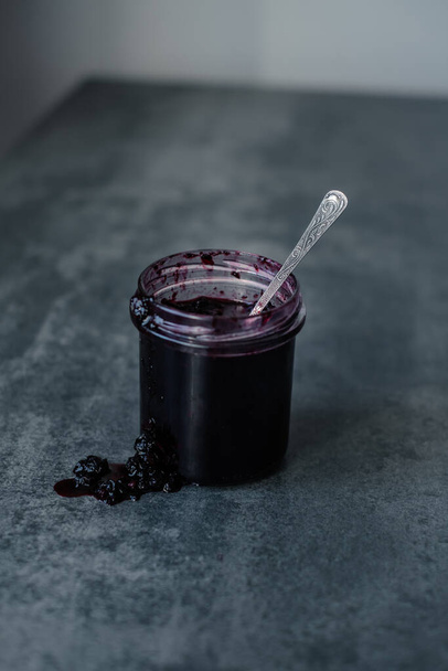 Abrir un frasco de mermelada de bayas con una cuchara en el centro. mermelada que fluye a lo largo de la pared de los frascos en la mesa. Foto de alta calidad - Foto, Imagen