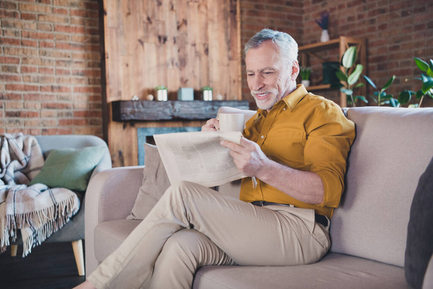 Photo of positive aged man sit on couch holding coffee reading interested newspaper enjoy home indoors - Zdjęcie, obraz