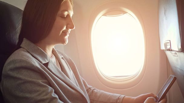 Brünette Frau schaut auf das Telefon, während sie am Flugzeugfenster sitzt - Foto, Bild