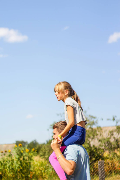 Defocused brother riding sister on back. Portrait of happy girl on man shoulders, piggyback. Girl fly. Family playing outside. Blue sky green tree background. Summer mood. Side view. Out of focus. - Fotografie, Obrázek
