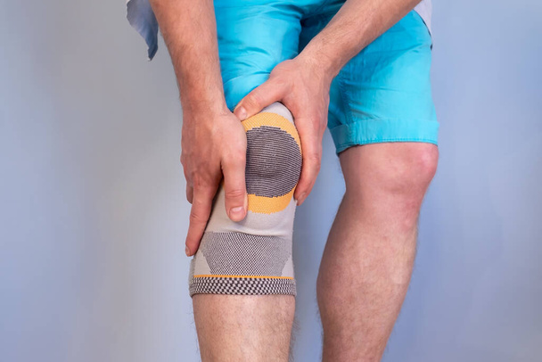 Articulación lesionada de la rodilla con un fijador especial que estabiliza el área del problema, con las manos masculinas sosteniendo la pierna lesionada. Fondo gris.  - Foto, Imagen