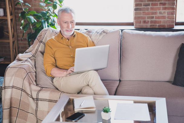 Portrait of positive aged man sit on sofa hold look use laptop watch video have good mood working from home indoors - Fotoğraf, Görsel