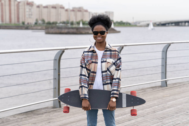 Junge afrikanische Mädchen Hipster lächelnd halten Longboard, lässige Skaterin im Freien über Flussblick - Foto, Bild