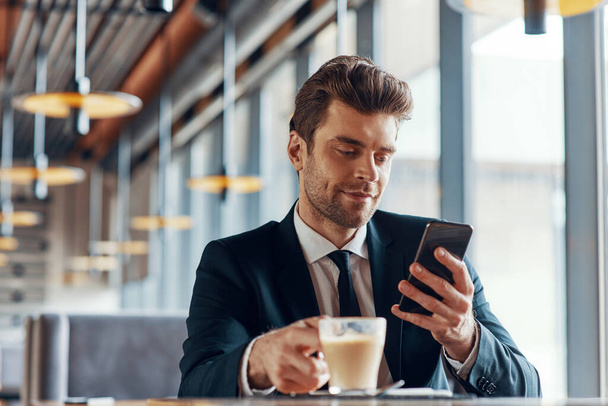 Handsome young man in full suit using smart phone and drinking coffee while sitting in the restaurant - 写真・画像