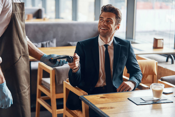 Handsome young smiling man in full suit making a contactless payment while sitting in the restaurant - 写真・画像
