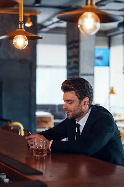 Handsome young man in full suit using smart phone while sitting at the bar counter in restaurant - Zdjęcie, obraz