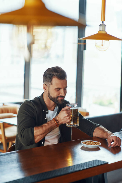 Handsome young man in casual clothing enjoying beer and smiling while spending time in the pub - 写真・画像