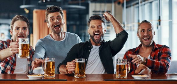 Cheering young men in casual clothing drinking beer and watching sport game - Фото, зображення