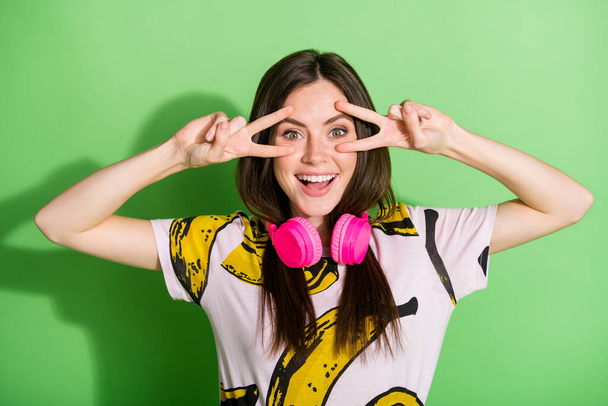 Photo of young happy excited cheerful girl in headphones showing v-sign on eyes isolated on green color background - Foto, immagini