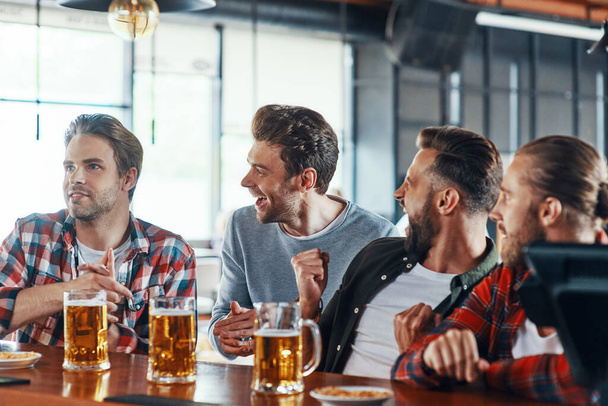 Cheering young men in casual clothing enjoying beer while sitting in the pub - Photo, Image