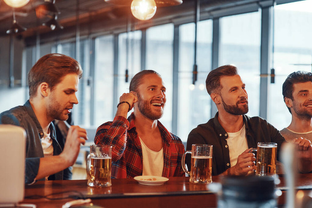Group of happy young men in casual clothing enjoying beer while sitting at the bar counter in pub - Фото, изображение