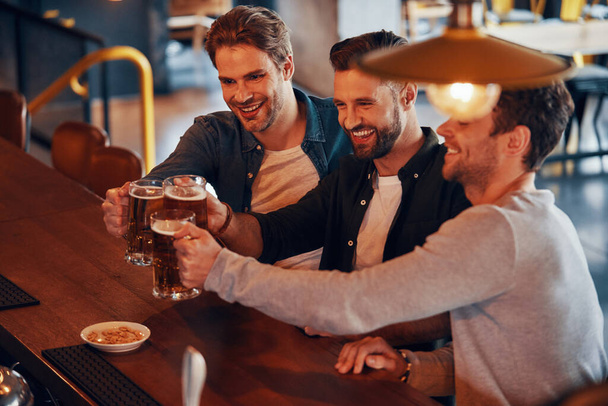 Top view of happy young men toasting each other with beer and smiling while sitting at the bar counter in pub - Фото, изображение