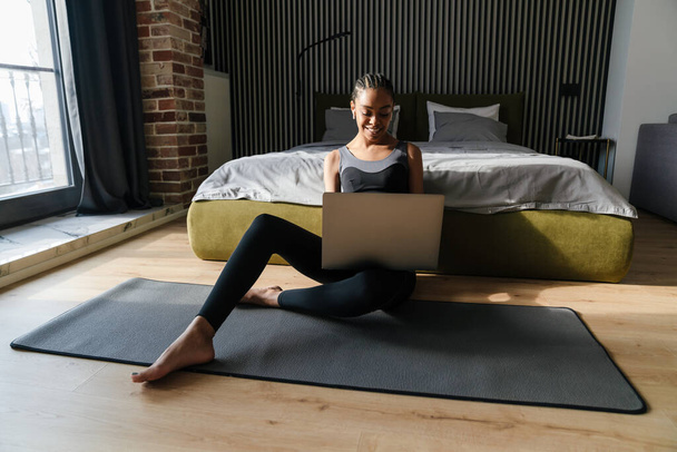Healthy smiling young fit african sportswoman using laptop computer while sitting on a fitness mat in the bedroom - 写真・画像
