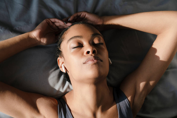 Relaxed african american girl listening music with earphones while lying on bed at home - 写真・画像