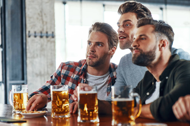 Three young men in casual clothing drinking beer and watching sport game while sitting in the pub - Фото, изображение