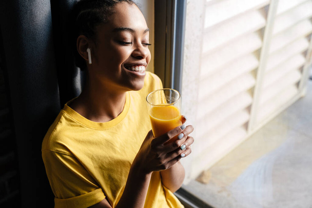 Pleased african american girl listening music with earphones while sitting at window in home, holding glass of orange juice - Foto, Imagen