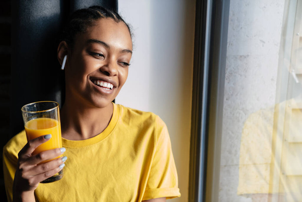 Задоволена афро-американська дівчинка, яка слухає музику з навушниками, сидячи біля вікна, тримає у руках склянку апельсинового соку. - Фото, зображення
