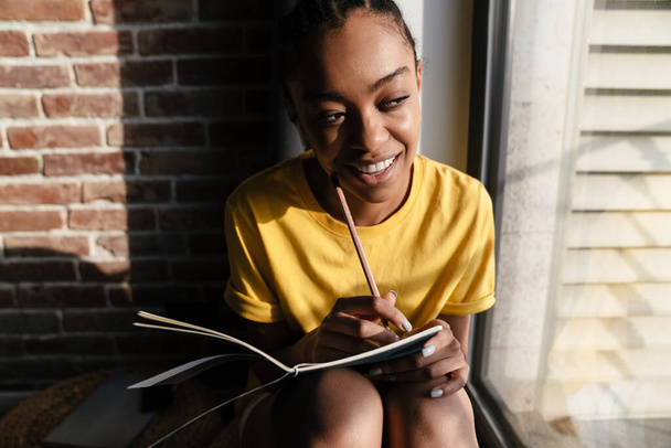 Happy african american girl in earphones writing down notes while sitting at window in home - Fotó, kép