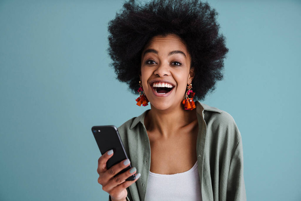 Happy african american girl smiling while using mobile phone isolated over blue background - Fotoğraf, Görsel