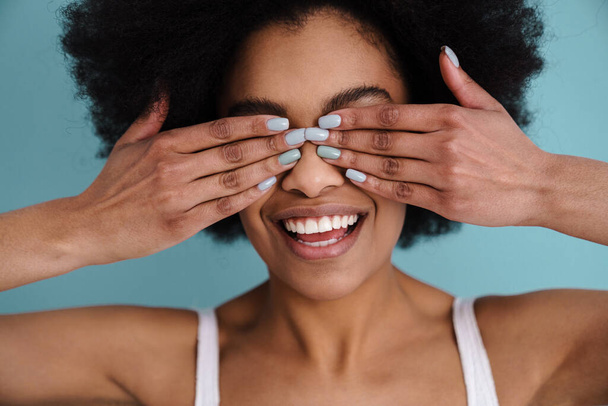 Funny african american girl smiling while covering her eyes isolated over blue background - Foto, Imagem