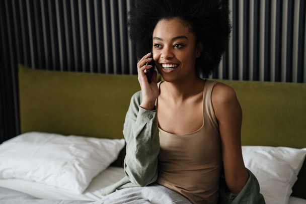 Happy african american girl talking on mobile phone while sitting in bed after sleep at home - Фото, зображення