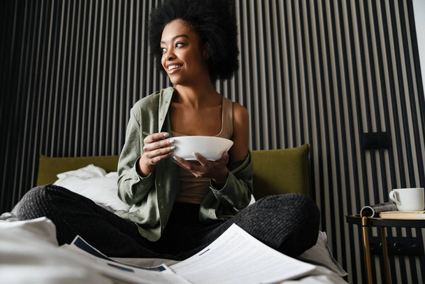 Happy african american girl examining documents while eating cereal in bed at home - Foto, Imagen