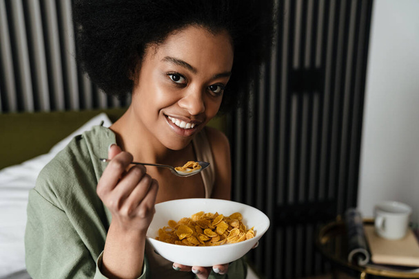 Smiling african american young woman eating breakfast while sitting in bed - Foto, Imagem