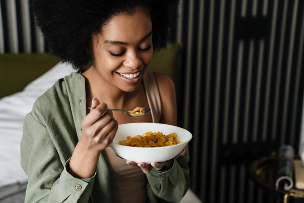 Smiling african american young woman eating breakfast while sitting in bed - Foto, immagini