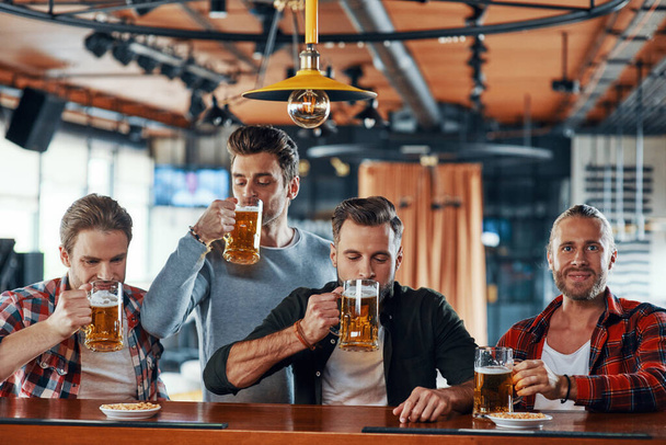 Group of cheerful young men in casual clothing enjoying beer while sitting at the bar counter in pub - Foto, Bild