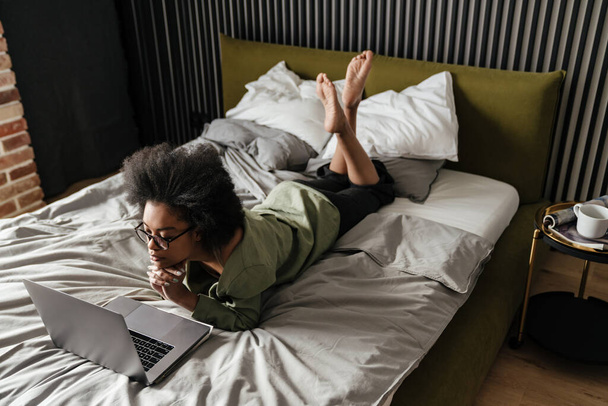 Mid aged african woman using laptop computer while laying in bed at home - 写真・画像