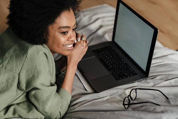 Happy Mid aged african woman using laptop computer while laying in bed at home - Фото, изображение