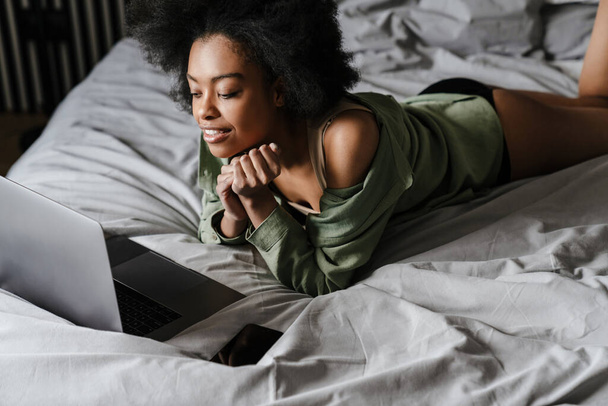 Mutlu Afrikalı Amerikalı kız evde uyuduktan sonra yatağında dizüstü bilgisayar kullanıyor. - Fotoğraf, Görsel