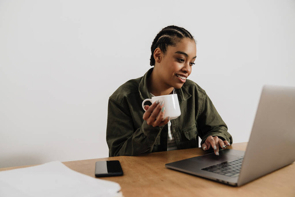 Black smiling woman drinking coffee while working with laptop in office - Photo, Image