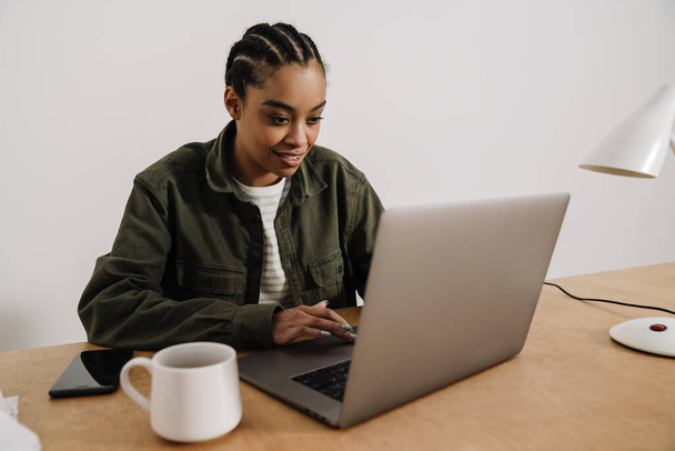 Black smiling woman working with laptop while sitting at table in office - Photo, Image