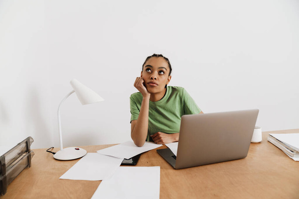 Black thinking woman working with laptop and papers while sitting at table in office - Foto, Imagem