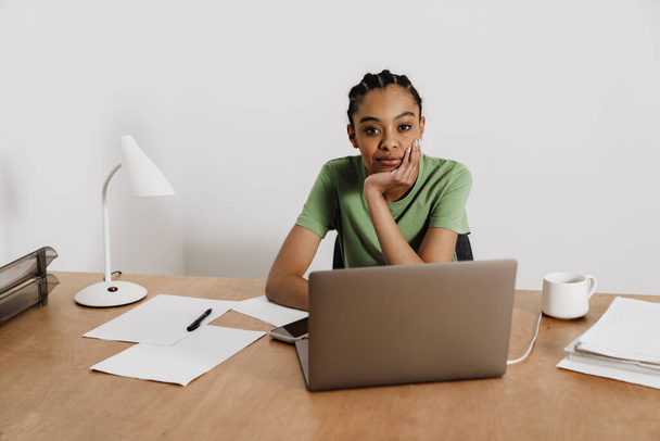 Czarna młoda kobieta pracuje z laptopem siedząc przy stole w biurze - Zdjęcie, obraz