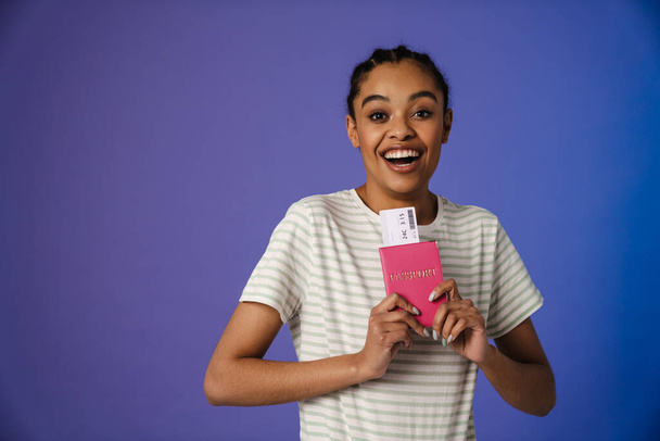 Excited black woman in t-shirt holding passport with ticket isolated over blue background - Foto, afbeelding