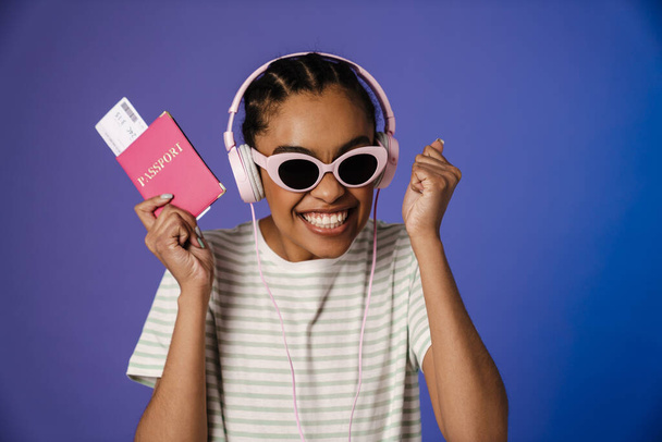 Excited black woman in headphones holding passport with ticket isolated over blue background - Foto, Imagen
