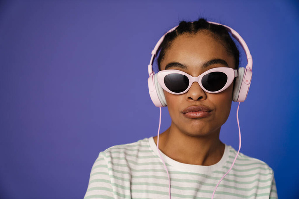 Young black woman in headphones posing at camera isolated over blue background - Φωτογραφία, εικόνα