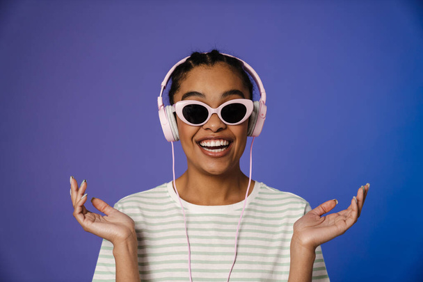Excited black woman in headphones gesturing and smiling at camera isolated over blue background - Valokuva, kuva
