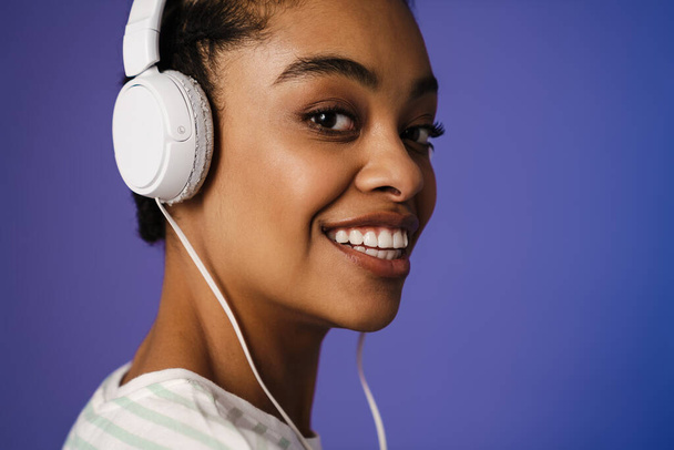 Close up of a smiling young african woman wearing headphones standing over blue wall - Foto, immagini