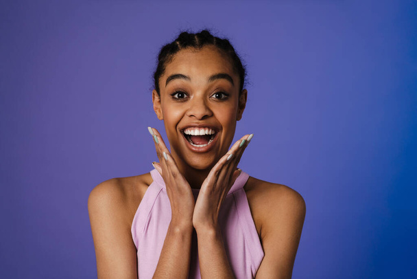 Happy young african woman standing over blue wall background, looking at camera with excitement - Zdjęcie, obraz