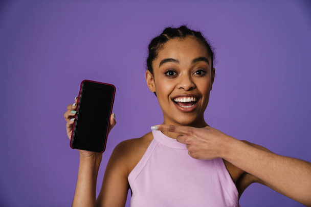 Excited black woman showing and pointing finger at mobile phone isolated over purple background - Foto, Imagen