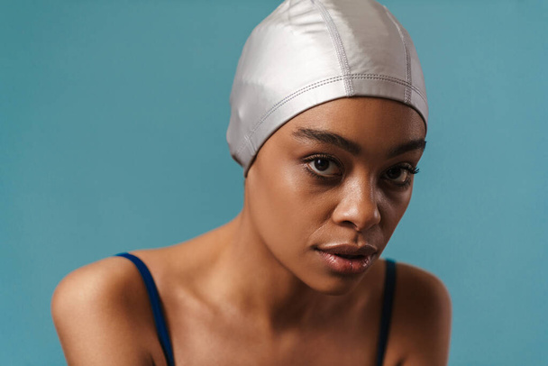 Close up portrait of a young african woman in swimsuit and swim hat standing over blue wall - Photo, Image